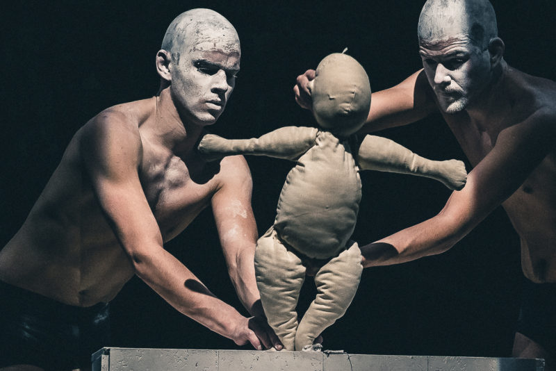 Two artists with white made-up heads steer a small puppet in the spotlight of a stage.