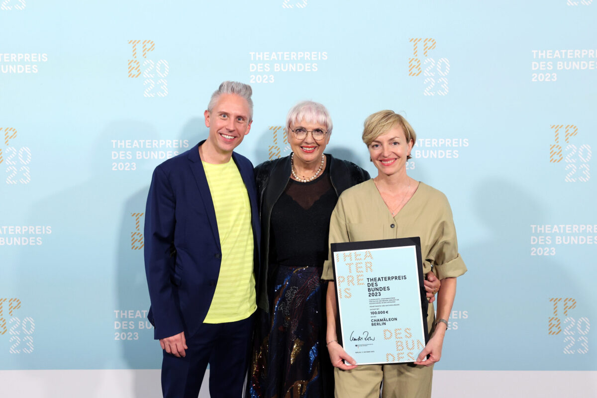 Geschäftsführer Hendrik Frobel und Intendantin Anke Politz stehen vor einer Fotowand und halten den Theaterpreis des Bundes. In ihrer Mitte steht Claudia Roth.