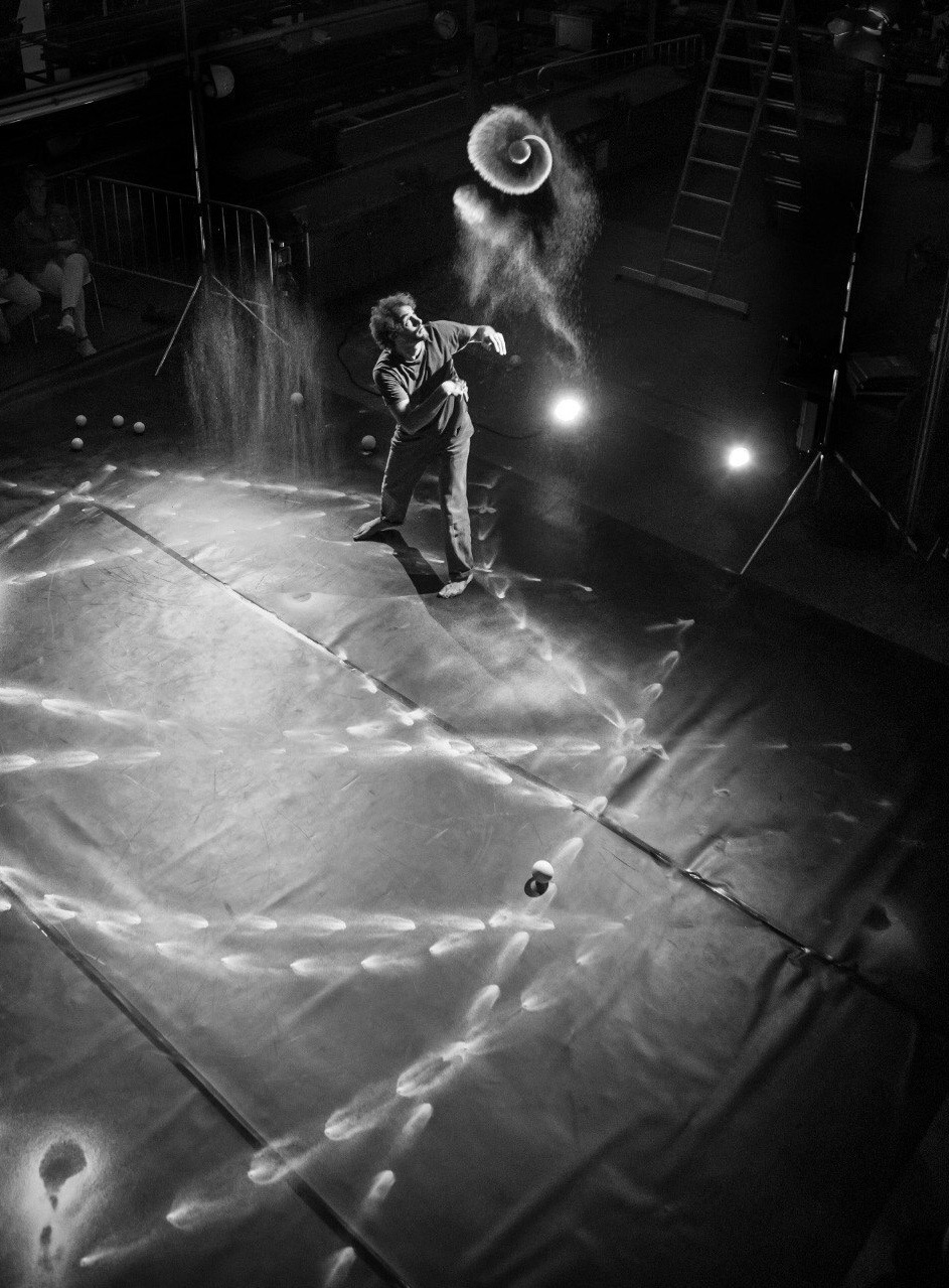 Tobias bei den Proben von seinem Stück Raum.Zeit. Er wirft einen Jonglierball in die Luft, der eine Spirale aus Sand nach sich zieht. Der Bühnenboden ist kreuz und quer mit Spuren aus Sand bedeckt.