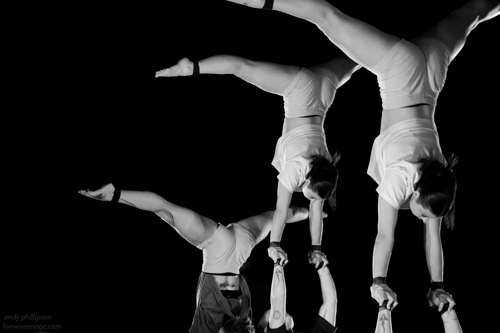Vor einem schwarzen Hintergrund machen Künstler*innen Hand auf Hand Akrobatik. In der Mitte ist eine weiblich gelesene Frau im Spagat-Handstand auf den Händen von einer anderen weiblich gelesenen Künstlerin. Das Bild dieser zwei Frauen ist in der rechten Bildseite dupliziert. In der linken Bildhälfte ist eine weitere weiblich gelesene Künstler*in im Spagat-Handstand auf den Händen einer weiteren Person.