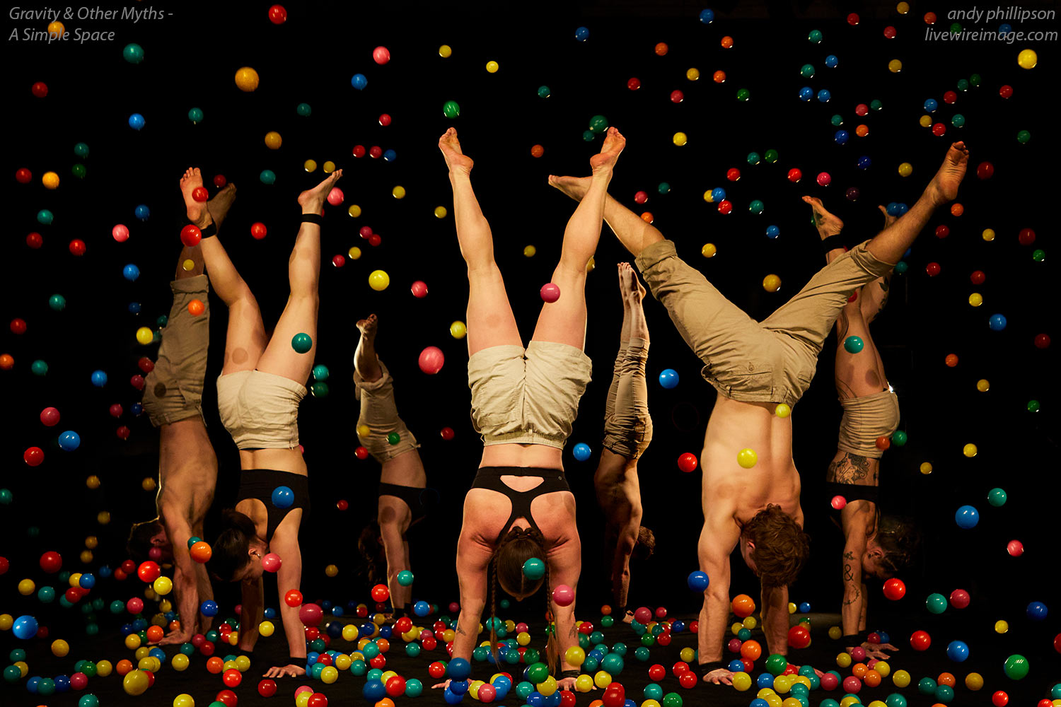 Showfotgrafie von A Simple Space. Die Künstler*innen stehen im Handstand im Kreis vor einem schwarzen Hintergrund. Viele bunte Bälle fliegen ums sie herum.