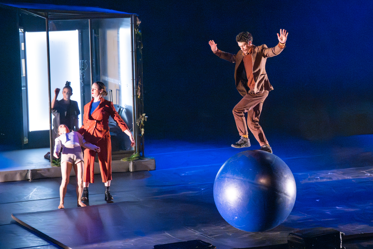From the show: a man is walking on a ball; three people in the background are doing somet…