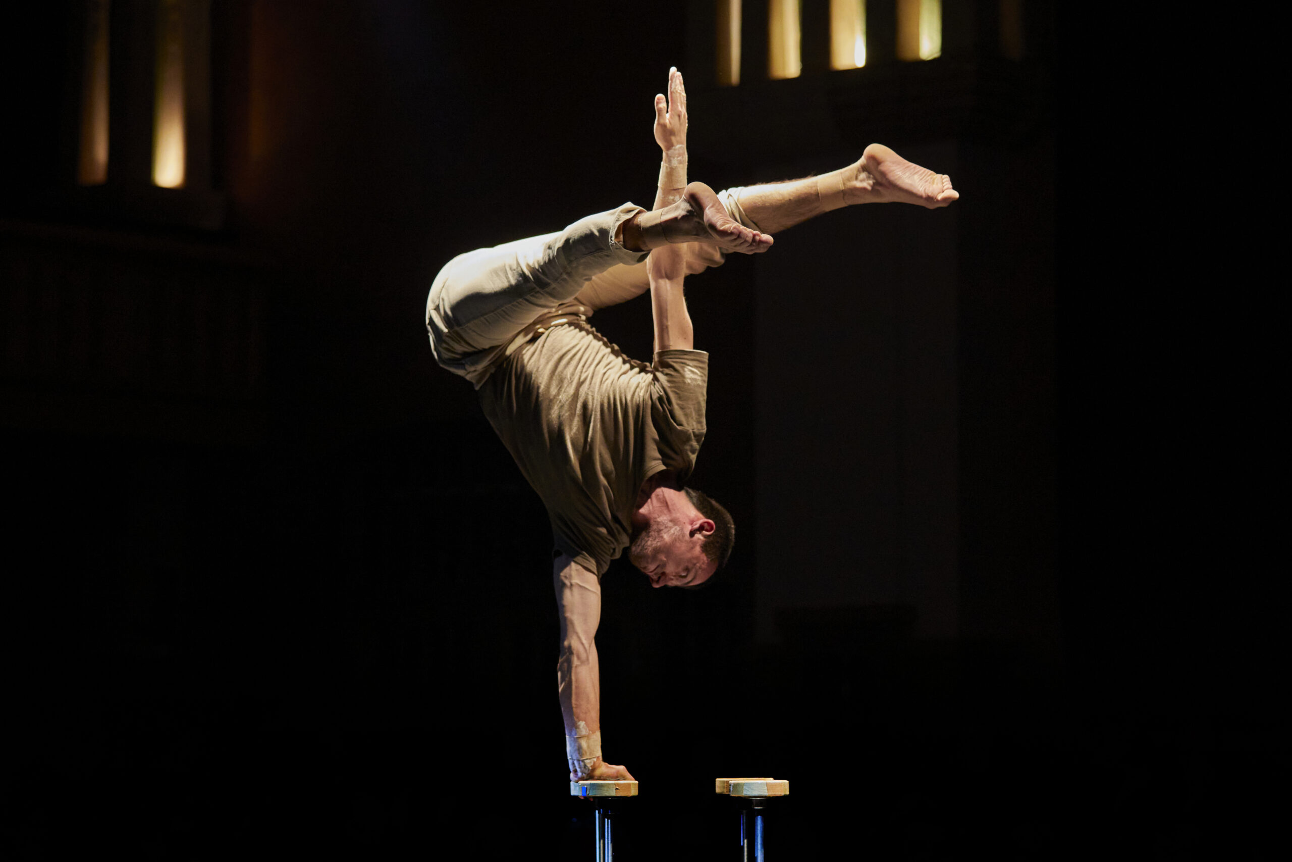 Aus dem Stück: Der Handstandkünstler Tony, macht einen einarmigen Handstand auf Stäben. Seine Beine befinden sich gegrätscht und im 90° Grad Winkel zu seinem Körper. 